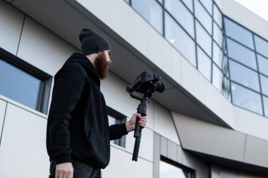 Siyah kapüşonlu sakallı profesyonel kameraman elinde 3 eksenli titreşim dengeleyicisi var. Film yapımcısı profesyonel bir sinema kamerasıyla harika bir video çekiyor. Görüntü Yönetmeni.