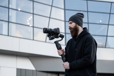 Siyah kapüşonlu sakallı profesyonel kameraman elinde 3 eksenli titreşim dengeleyicisi var. Film yapımcısı profesyonel bir sinema kamerasıyla harika bir video çekiyor. Görüntü Yönetmeni.
