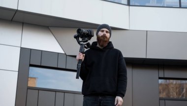 Siyah kapüşonlu sakallı profesyonel kameraman elinde 3 eksenli titreşim dengeleyicisi var. Film yapımcısı profesyonel bir sinema kamerasıyla harika bir video çekiyor. Görüntü Yönetmeni.