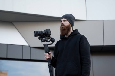 Siyah kapüşonlu sakallı profesyonel kameraman elinde 3 eksenli titreşim dengeleyicisi var. Film yapımcısı profesyonel bir sinema kamerasıyla harika bir video çekiyor. Görüntü Yönetmeni.