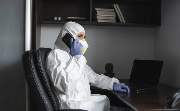 El hombre con traje protector, máscara médica y guantes de goma se sienta en casa y habla por teléfono con amigos o compañeros de negocios durante la cuarentena. Un hombre trabajando a distancia en un covidio pandémico. Coronavirus . — Foto de Stock