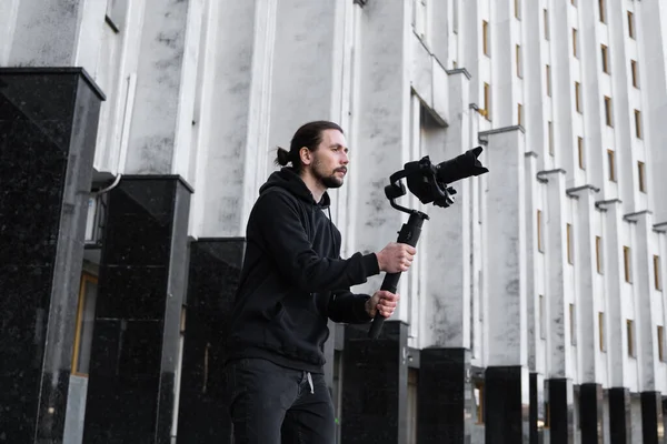 Jonge professionele videograaf met professionele camera op 3-assige gimbal stabilisator. Pro-apparatuur helpt om video van hoge kwaliteit te maken zonder te schudden. Cameraman dragen zwarte hoodie het maken van een video 's. — Stockfoto