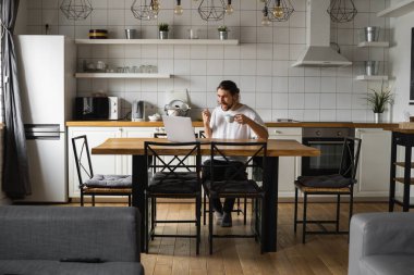 Genç serbest çalışan, projesini evden yürütüyor. Genç serbest çalışanın yumruklarıyla zafer kazanmasına sevindim. Mutluluk kazanan mutlu adam evdeki masada dizüstü bilgisayar kullanıyor. Başarılı sakallı adam..