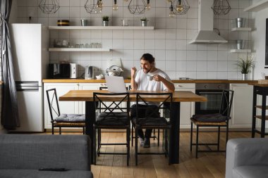 Genç serbest çalışan, projesini evden yürütüyor. Genç serbest çalışanın yumruklarıyla zafer kazanmasına sevindim. Mutluluk kazanan mutlu adam evdeki masada dizüstü bilgisayar kullanıyor. Başarılı sakallı adam..