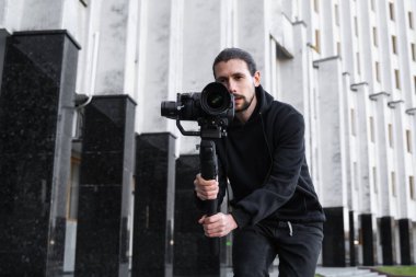 3 eksenli titreşimli dengeleyicide profesyonel kamera tutan genç profesyonel kameraman. Pro ekipmanlar titremeden yüksek kaliteli video çekmeye yardımcı olur. Siyah kapüşonlu bir kameraman video çekiyor..