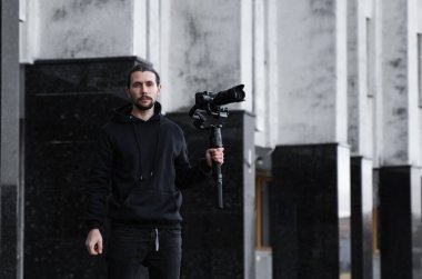 3 eksenli titreşimli dengeleyicide profesyonel kamera tutan genç profesyonel kameraman. Pro ekipmanlar titremeden yüksek kaliteli video çekmeye yardımcı olur. Siyah kapüşonlu bir kameraman video çekiyor..