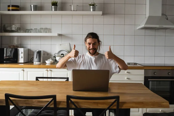 兴奋的商人对成功的项目感到高兴.男人把大拇指举到空中，坐在厨房里看着笔记本电脑。快乐的自由职业者，在家里笔记本电脑上工作的学生. — 图库照片