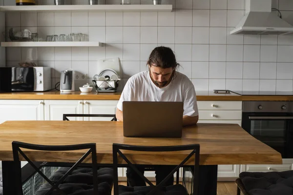 自由撰稿人在家里厨房里工作，使用笔记本电脑。带着胡子的男人一边用笔记本电脑一边读坏消息。英俊的成功的自我企业家坐在他现代的家里工作. — 图库照片