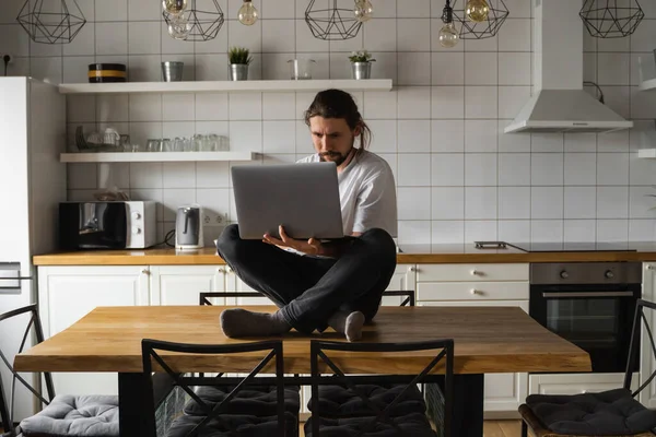 Ελεύθερος επαγγελματίας που εργάζεται από το σπίτι κάθεται στην κορυφή του τραπεζιού της κουζίνας και χρησιμοποιώντας το laptop. Γενειοφόρος που δουλεύει με λάπτοπ και διαβάζει ειδήσεις. Επιτυχημένος επιχειρηματίας που εργάζεται στο σύγχρονο σπίτι του. — Φωτογραφία Αρχείου