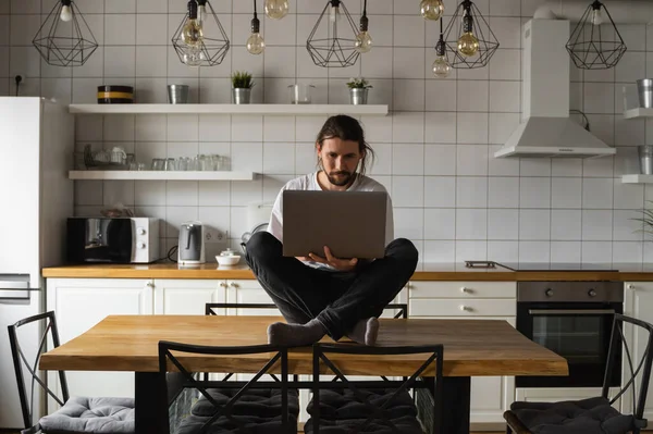 Ελεύθερος επαγγελματίας που εργάζεται από το σπίτι κάθεται στην κορυφή του τραπεζιού της κουζίνας και χρησιμοποιώντας το laptop. Γενειοφόρος που δουλεύει με λάπτοπ και διαβάζει ειδήσεις. Επιτυχημένος επιχειρηματίας που εργάζεται στο σύγχρονο σπίτι του. — Φωτογραφία Αρχείου