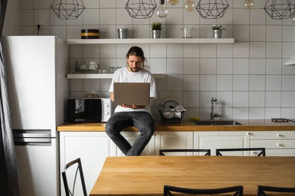 自由职业者坐在厨房的工作台上，使用笔记本电脑，从家里开始工作。带着胡子的男人在用笔记本电脑和阅读新闻。在他现代家里工作的漂亮的成功的自我企业家. — 图库照片