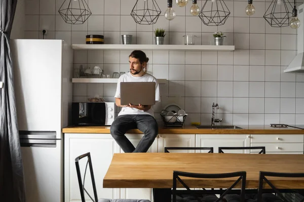 Ελεύθερος επαγγελματίας που εργάζεται από το σπίτι κάθεται σε έναν πάγκο κουζίνας και χρησιμοποιώντας το lap-top. Γενειοφόρος που δουλεύει με λάπτοπ και διαβάζει ειδήσεις. Όμορφος επιτυχημένος επιχειρηματίας που εργάζεται στο σύγχρονο σπίτι του. — Φωτογραφία Αρχείου
