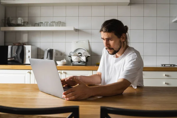 自由撰稿人在家里厨房里工作，使用笔记本电脑。带着胡子的男人用笔记本电脑工作，阅读好消息。英俊的成功的自我企业家坐在他现代的家里工作. — 图库照片