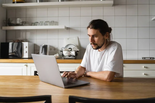 自由撰稿人在家里厨房里工作，使用笔记本电脑。带着胡子的男人用笔记本电脑工作，阅读好消息。英俊的成功的自我企业家坐在他现代的家里工作. — 图库照片
