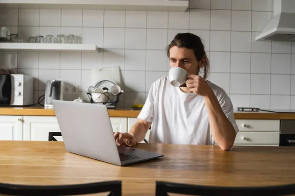 自由撰稿人在家里厨房里工作，使用笔记本电脑。英俊的面带微笑的男人端着一杯咖啡，看着笔记本电脑。成功的自我创业者，坐在笔记本电脑旁工作. — 图库照片