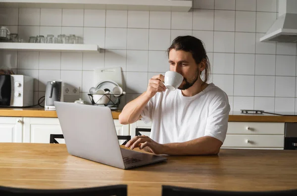 Ελεύθερος επαγγελματίας που εργάζεται από το σπίτι στην κουζίνα και χρησιμοποιώντας το lap-top. Όμορφος χαμογελαστός άντρας που κρατάει ένα φλιτζάνι καφέ και κοιτάει το λάπτοπ. Επιτυχημένος επιχειρηματίας κάθεται με φορητό υπολογιστή και εργασίας. — Φωτογραφία Αρχείου