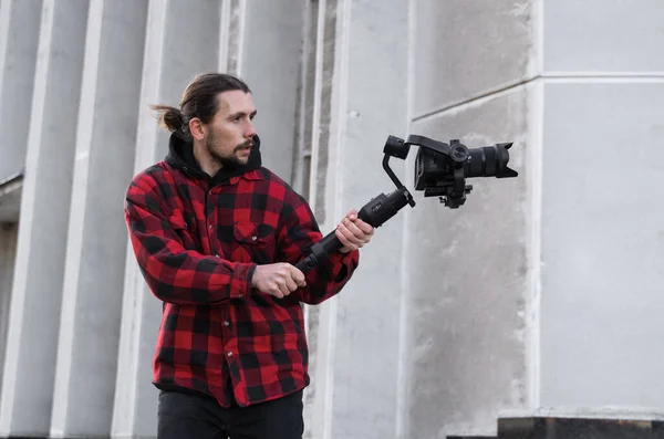 Giovane videografo professionale con fotocamera professionale su stabilizzatore gimbale a 3 assi. L'attrezzatura Pro aiuta a fare video di alta qualità senza scuotere. Cameraman indossa camicia rossa facendo un video . — Foto Stock