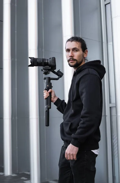 Giovane videografo professionale con fotocamera professionale su stabilizzatore gimbale a 3 assi. L'attrezzatura Pro aiuta a fare video di alta qualità senza scuotere. Cameraman indossa felpa bianca facendo un video . — Foto Stock