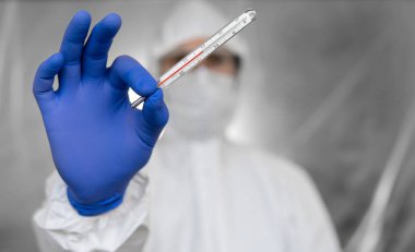 Beyaz koruyucu giysili doktor, tıbbi maske ve lastik eldivenler koronavirüs salgınında termometre tutuyor. Salgın, Coronavirus covid 19 salgını. Sıcaklık ölçülüyor ve gösteriliyor.