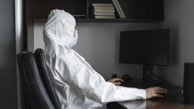 Tıbbi maskeli ve koruyucu giysili bir adam, lastik eldivenler evde oturuyor ve karantina sırasında masada bilgisayarla çalışıyor. Tasarımcı, sanatçı, mimar, iş adamı salgın bir covid 'de uzaktan çalışıyor..