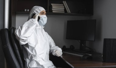 Koruyucu takım elbiseli, tıbbi maskeli ve lastik eldivenli bir adam evde oturuyor ve karantina sırasında arkadaşlarıyla veya iş ortağıyla telefonda konuşuyor. Uzaktan kumandalı bir adam salgın hastalığın pençesinde. Coronavirüs.
