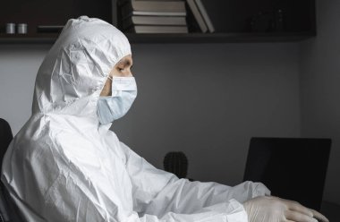 Koruyucu giysili, tıbbi maskeli ve lastik eldivenli bir adam evde oturuyor ve karantina sırasında masada dizüstü bilgisayarla çalışıyor. İnsan, tasarımcı, sanatçı, mimar, bir salgında uzaktan çalışan iş adamı