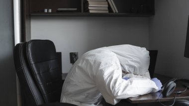 Tıbbi maskeli ve koruyucu giysili bir adam, eldivenler evde oturuyor ve kendini kötü hissediyor. Karantina sırasında masada dizüstü bilgisayarla çalışıyor. Bir salgın covid içinde uzaktan çalışma.
