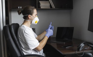 Tıbbi maskeli ve koruyucu giysili bir adam, eldivenler evde oturuyor ve karantina sırasında masada bilgisayarla çalışıyor ve başparmağını kaldırıyor. Bir salgın covid içinde uzaktan çalışma.