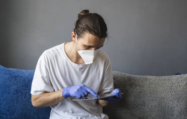Beyaz gömlekli, tıbbi maskeli ve lastik eldivenli bir adam evde oturuyor ve karantina sırasında kanepede tabletle çalışıyor. İnsan, tasarımcı, sanatçı, mimar, iş adamı salgın bir covid 'de uzaktan çalışıyor.