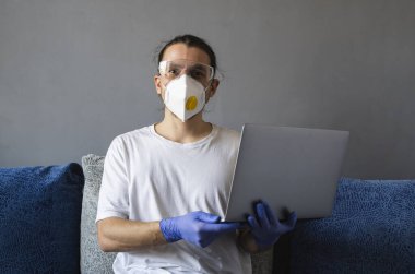 Tıbbi maskeli ve lastik eldivenli bir adam evde oturuyor ve karantina sırasında kanepede dizüstü bilgisayarla çalışıyor. Tasarımcı, sanatçı, mimar, salgın bir covid 'in uzaktan kumandalı işadamı. Coronavirüs.