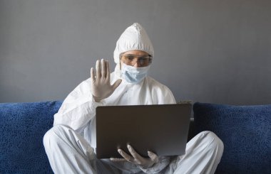 Koruyucu giysili, tıbbi maskeli ve lastik eldivenli bir adam evde oturuyor ve karantina sırasında masada dizüstü bilgisayarla çalışıyor. Tasarımcı, sanatçı, mimar, iş adamı salgın bir covid 'de uzaktan çalışıyor..