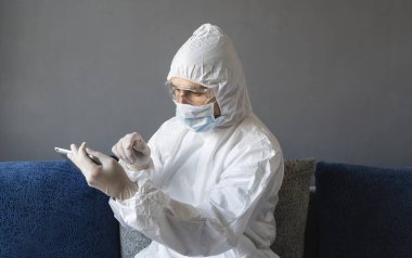 Koruyucu giysili, tıbbi maskeli ve lastik eldivenli bir adam evde oturuyor ve karantina sırasında kanepede tabletle çalışıyor. Tasarımcı, sanatçı, mimar, iş adamı salgın bir covid 'de uzaktan çalışıyor..