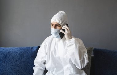 Koruyucu takım elbiseli, tıbbi maskeli ve lastik eldivenli bir adam evde oturuyor ve karantina sırasında bir kanepede arkadaşlarıyla veya iş arkadaşlarıyla konuşuyor. Salgın bir covid 'de uzaktan çalışan bir adam..