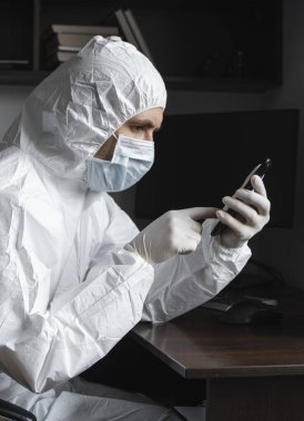 Koruyucu giysili, tıbbi maskeli ve lastik eldivenli bir adam evde oturuyor ve karantina sırasında arkadaşlarıyla veya iş ortaklarıyla sohbet etmek için telefon kullanıyor. Salgın bir covid 'de uzaktan çalışan bir adam.