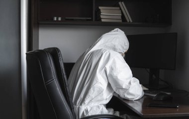 Tıbbi maskeli ve koruyucu giysili bir adam, lastik eldivenler evde oturuyor ve karantina sırasında masada bilgisayarla çalışıyor. Tasarımcı, sanatçı, mimar, iş adamı salgın bir covid 'de uzaktan çalışıyor..