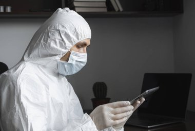 Koruyucu giysili, tıbbi maskeli ve lastik eldivenli bir adam evde oturuyor ve karantina sırasında masada tabletle çalışıyor. Tasarımcı, sanatçı, mimar, iş adamı salgın bir covid 'de uzaktan çalışıyor..