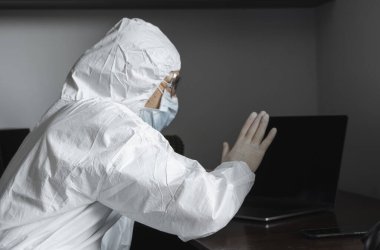 Tıbbi maskeli ve koruyucu elbiseli bir adam, eldivenler evde oturuyor ve karantina sırasında laptopta çalışıyor ve iş ortağına ya da arkadaşlarına merhaba diyor. Bir salgın covid içinde uzaktan çalışma.