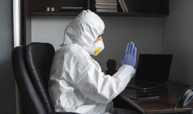 Tıbbi maskeli ve koruyucu elbiseli bir adam, eldivenler evde oturuyor ve karantina sırasında laptopta çalışıyor ve iş ortağına ya da arkadaşlarına merhaba diyor. Bir salgın covid içinde uzaktan çalışma.