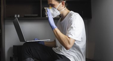 Tıbbi maskeli ve lastik eldivenli bir adam evde masada oturuyor ve karantina sırasında dizüstü bilgisayarla çalışıyor. Tasarımcı, sanatçı, mimar, salgın bir covid 'in uzaktan kumandalı işadamı. Coronavirüs.