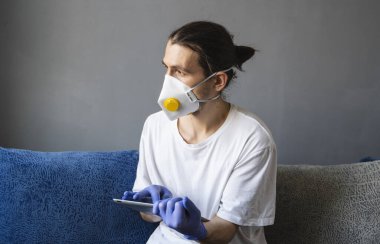 Beyaz gömlekli, tıbbi maskeli ve lastik eldivenli bir adam evde oturuyor ve karantina sırasında kanepede tabletle çalışıyor. İnsan, tasarımcı, sanatçı, mimar, iş adamı salgın bir covid 'de uzaktan çalışıyor.
