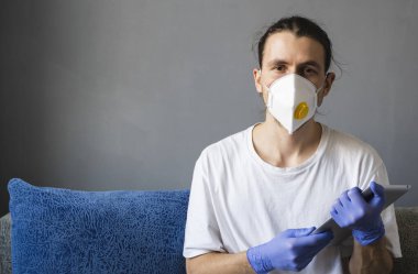 Beyaz gömlekli, tıbbi maskeli ve lastik eldivenli bir adam evde oturuyor ve karantina sırasında kanepede tabletle çalışıyor. İnsan, tasarımcı, sanatçı, mimar, iş adamı salgın bir covid 'de uzaktan çalışıyor.