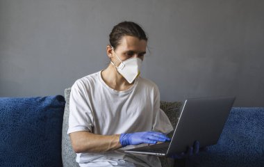 Tıbbi maskeli ve lastik eldivenli bir adam evde oturuyor ve karantina sırasında kanepede dizüstü bilgisayarla çalışıyor. Tasarımcı, sanatçı, mimar, salgın bir covid 'in uzaktan kumandalı işadamı. Coronavirüs.