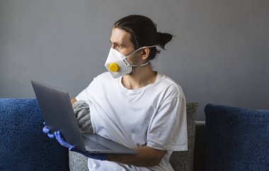 Tıbbi maskeli ve lastik eldivenli bir adam evde oturuyor ve karantina sırasında kanepede dizüstü bilgisayarla çalışıyor. Tasarımcı, sanatçı, mimar, salgın bir covid 'in uzaktan kumandalı işadamı. Coronavirüs.