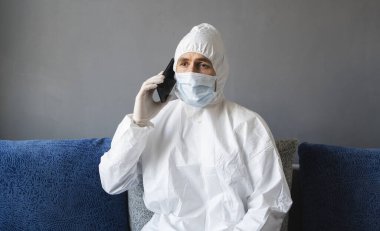 Koruyucu takım elbiseli, tıbbi maskeli ve lastik eldivenli bir adam evde oturuyor ve karantina sırasında bir kanepede arkadaşlarıyla veya iş arkadaşlarıyla konuşuyor. Salgın bir covid 'de uzaktan çalışan bir adam..