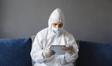 Koruyucu giysili, tıbbi maskeli ve lastik eldivenli bir adam evde oturuyor ve karantina sırasında kanepede tabletle çalışıyor. Tasarımcı, sanatçı, mimar, iş adamı salgın bir covid 'de uzaktan çalışıyor..