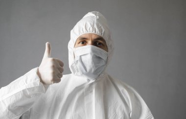 Beyaz koruyucu giysili, tıbbi maskeli ve lastik eldivenli doktor baş parmağını kaldırıyor. Koronavirüs salgını tehdidi altındayken insanlara yardım etmek. Salgın, Coronavirus covid 19 salgını..