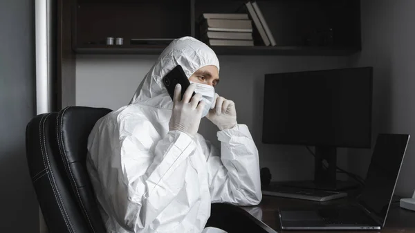 Egy férfi védőruhában, orvosi maszkban és gumikesztyűben otthon ül, és telefonon beszél barátaival vagy üzlettársával a karantén alatt. Egy ember a távoli munkában, egy járvány idején. Koronavírus. — Stock Fotó
