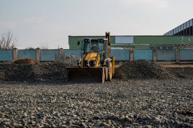 İnşaat alanında çakıllı çalışan sarı tekerlek doldurucu kazıyıcı makinesi. Asfaltlama için temel hazırlıklar yapılıyor. Yol inşaatı. Park binası..