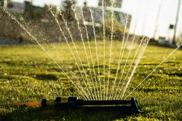 Разбрызгиватель лужайки брызгает водой зеленую траву. Современное устройство орошения садовой травы. Система орошения - методика полива в саду. Полив лужайки дождевателем . — стоковое фото