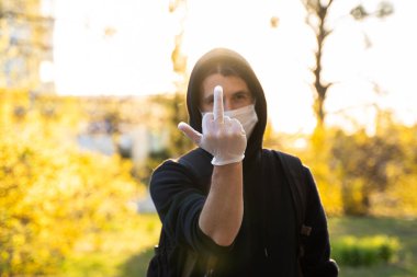 Siyah kapüşonlu bir adam orta parmağını gösteren tıbbi bir maske takıyor. Bakteriyel Coronavirus ya da Covid 19 salgınının önlenmesi. Koronavirüs küresel karantina.